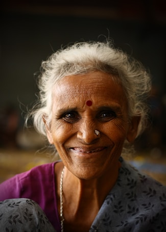 An elderly woman with gray hair and a joyful expression, wearing a colorful saree and traditional jewelry, including a nose ring and a red bindi on her forehead. The background is dark and blurred, drawing focus to her smiling face.