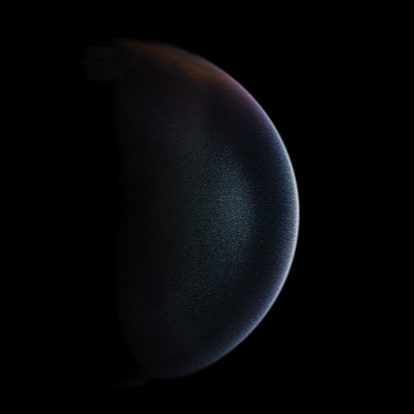 A spherical object with a dark surface is faintly illuminated on one side, against a completely black background, giving it a mysterious, otherworldly appearance.