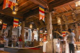 A richly decorated interior with intricate wooden beams and columns. Colorful flags hang from the ceiling, adding vibrancy to the setting. Several people dressed in traditional attire are engaged in a ceremonial procession, with instruments and ornate decorations visible. Warm lighting from ornate lamps creates a welcoming and spiritual ambiance.