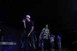 Several people are on stage, with one person singing into a microphone while wearing a white cap and black shirt. Another person next to him is singing energetically, wearing a floral shirt. A keyboard is visible on the left, and a few more people are singing or playing instruments further in the background.