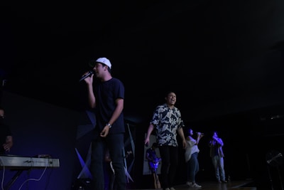 Several people are on stage, with one person singing into a microphone while wearing a white cap and black shirt. Another person next to him is singing energetically, wearing a floral shirt. A keyboard is visible on the left, and a few more people are singing or playing instruments further in the background.
