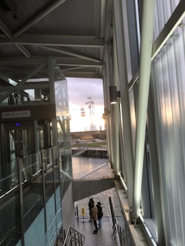 Inside a modern building with large glass panels and metal beams, there is an elevator shaft to the left, next to a glass wall. In the distance, cable cars are visible against a sunset. Below, a few people are standing near a body of water beside the building.