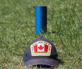 A cap with a patch featuring the Canadian flag and the words 'All But The Flag' and 'One Voice Speaks For Many' is placed on grass. Behind it stands a blue cylindrical object.