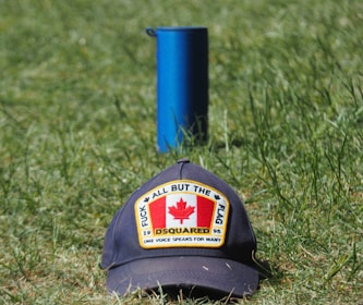 A cap with a patch featuring the Canadian flag and the words 'All But The Flag' and 'One Voice Speaks For Many' is placed on grass. Behind it stands a blue cylindrical object.