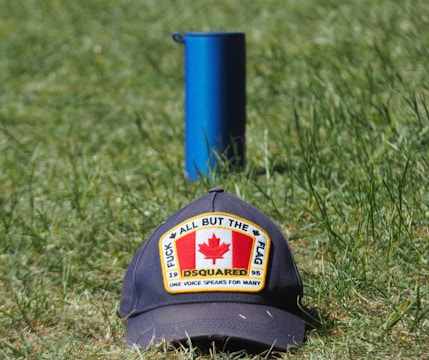 A cap with a patch featuring the Canadian flag and the words 'All But The Flag' and 'One Voice Speaks For Many' is placed on grass. Behind it stands a blue cylindrical object.