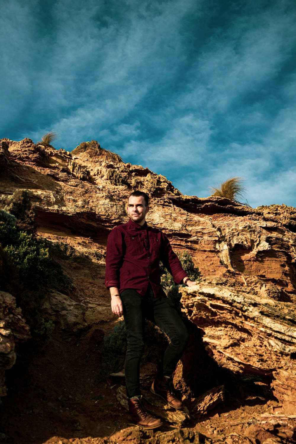 man in maroon long sleeve shirt and black pants sitting on brown rock formation under blue