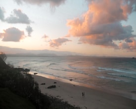 The image captures a serene beach scene at sunset. The foreground features a sandy beach with gentle waves lapping the shore. Two silhouetted figures stroll along the sand, adding a sense of tranquility and companionship. The sky is painted with soft hues of pink and orange, with scattered fluffy clouds enhancing the tranquil atmosphere.