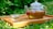 A glass teapot filled with herbal tea featuring red goji berries, placed on a wooden tray. The teapot is accompanied by a small, transparent glass cup. The scene is set outdoors amidst lush green foliage, suggesting a serene and natural setting.