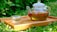 A glass teapot filled with herbal tea featuring red goji berries, placed on a wooden tray. The teapot is accompanied by a small, transparent glass cup. The scene is set outdoors amidst lush green foliage, suggesting a serene and natural setting.
