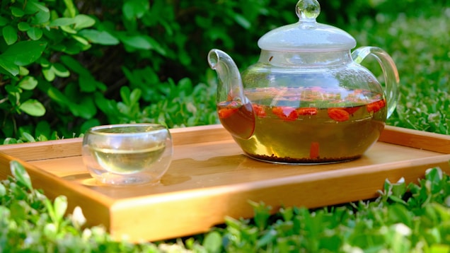 A glass teapot filled with herbal tea featuring red goji berries, placed on a wooden tray. The teapot is accompanied by a small, transparent glass cup. The scene is set outdoors amidst lush green foliage, suggesting a serene and natural setting.