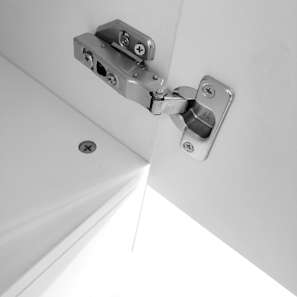 A close-up of a metal hinge attached to a white cabinet door. The hinge is secured with screws and is in an open position, exposing the edge of the cabinet and the door's interior.