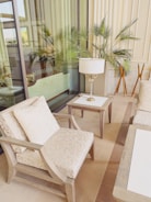 The image depicts a well-furnished balcony or patio area featuring a cushioned armchair with floral patterns and a square side table. A decorative lamp is placed on the table. Large glass doors reflect the interior, showing a green sofa inside. Potted palm plants add a touch of greenery, and several folded wooden easels or stands are visible in the background.