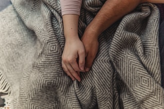 Two hands are gently holding each other on top of a patterned, grey and white blanket, creating a sense of connection and warmth.