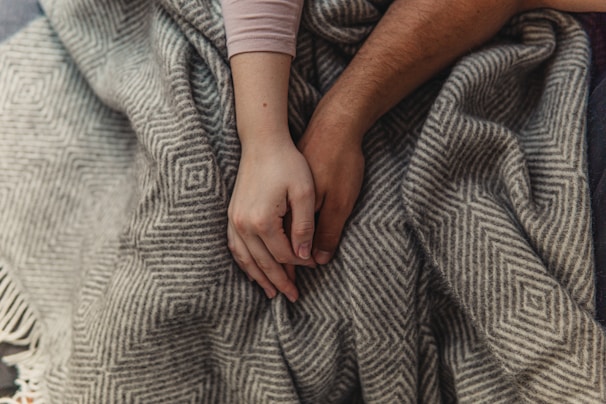 Two hands are gently holding each other on top of a patterned, grey and white blanket, creating a sense of connection and warmth.