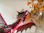 A detailed, vibrant sculpture of a dragon featured. Its wings are extended, showing intricate vein patterns, and its mouth is open in a fierce expression. In the background, decorative dried leaves or flowers in shades of gold and brown are partially visible.