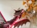 A detailed, vibrant sculpture of a dragon with red and maroon coloring is prominently featured. Its wings are extended, showing intricate vein patterns, and its mouth is open in a fierce expression. In the background, decorative dried leaves or flowers in shades of gold and brown are partially visible.