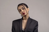 A person is wearing a black blazer with minimal makeup and bright red lipstick, posing against a plain gray background. The hair is neatly tied back with a few strands loosely hanging. The attire and posture suggest a formal or professional setting.