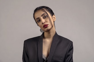 A person is wearing a black blazer with minimal makeup and bright red lipstick, posing against a plain gray background. The hair is neatly tied back with a few strands loosely hanging. The attire and posture suggest a formal or professional setting.