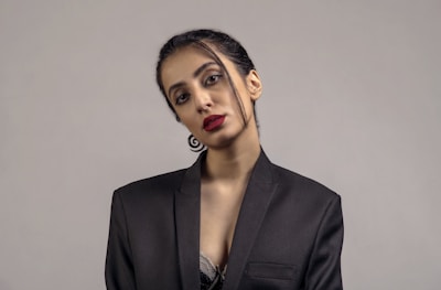 A person is wearing a black blazer with minimal makeup and bright red lipstick, posing against a plain gray background. The hair is neatly tied back with a few strands loosely hanging. The attire and posture suggest a formal or professional setting.