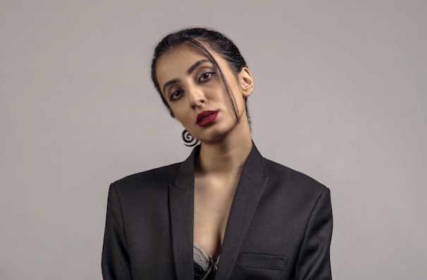 A person is wearing a black blazer with minimal makeup and bright red lipstick, posing against a plain gray background. The hair is neatly tied back with a few strands loosely hanging. The attire and posture suggest a formal or professional setting.