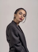 A person posing against a plain background, dressed in a formal black blazer. The individual has neatly styled dark hair with an elegant updo and wears red lipstick, adding a touch of sophistication. The expression is poised and confident.