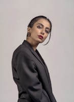 A person posing against a plain background, dressed in a formal black blazer. The individual has neatly styled dark hair with an elegant updo and wears red lipstick, adding a touch of sophistication. The expression is poised and confident.