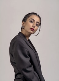A person posing against a plain background, dressed in a formal black blazer. The individual has neatly styled dark hair with an elegant updo and wears red lipstick, adding a touch of sophistication. The expression is poised and confident.