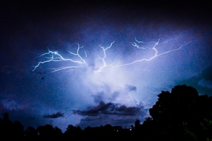 Bright flashes of lightning illuminate a dark, cloudy night sky. The lightning branches out in jagged patterns, casting an eerie glow over silhouetted trees below. The scene conveys a sense of power and raw energy in the midst of a storm.
