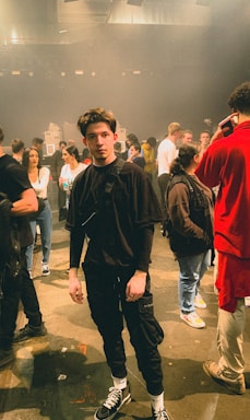 A group of people standing in a dimly lit indoor venue. The floor is concrete, and there are stage lights and speakers visible in the background. Individuals are casually dressed, appearing as if they are either waiting for an event to start or taking a break.