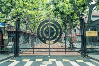 An ornate black metal gate with circular patterns stands closed in front of a pathway lined with trees. On either side are buildings with gray walls and red-tiled roofs. The area is shaded by tall green trees, casting a tranquil and inviting atmosphere. Signs with text in another language are visible on each side of the gate.