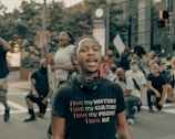 A group of people are gathered outdoors, some kneeling, and others standing. One person in the foreground wears a black t-shirt with the text 'I love my HISTORY, I love my CULTURE, I love my PEOPLE, I love ME.' The atmosphere seems active, with visible signs being held by participants.