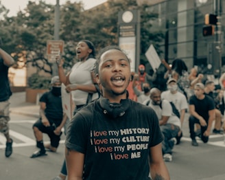 A group of people are gathered outdoors, some kneeling, and others standing. One person in the foreground wears a black t-shirt with the text 'I love my HISTORY, I love my CULTURE, I love my PEOPLE, I love ME.' The atmosphere seems active, with visible signs being held by participants.