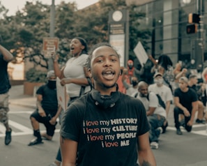 A group of people are gathered outdoors, some kneeling, and others standing. One person in the foreground wears a black t-shirt with the text 'I love my HISTORY, I love my CULTURE, I love my PEOPLE, I love ME.' The atmosphere seems active, with visible signs being held by participants.