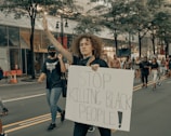 A group of people are participating in a protest march along a city street. The central figure, a woman with curly hair, holds up a sign that reads 'STOP KILLING BLACK PEOPLE!' and raises one hand. She is accompanied by other protesters, some wearing masks, walking in the same direction. Trees and commercial buildings line the street.