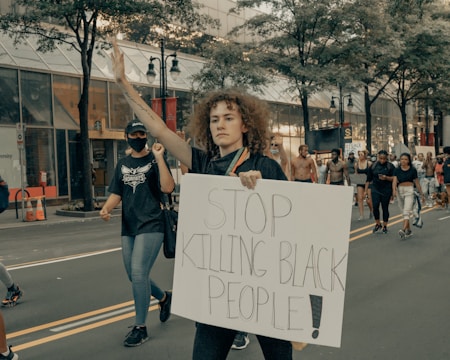 A group of people are participating in a protest march along a city street. The central figure, a woman with curly hair, holds up a sign that reads 'STOP KILLING BLACK PEOPLE!' and raises one hand. She is accompanied by other protesters, some wearing masks, walking in the same direction. Trees and commercial buildings line the street.