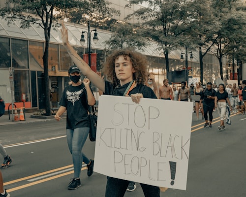 A group of people are participating in a protest march along a city street. The central figure, a woman with curly hair, holds up a sign that reads 'STOP KILLING BLACK PEOPLE!' and raises one hand. She is accompanied by other protesters, some wearing masks, walking in the same direction. Trees and commercial buildings line the street.