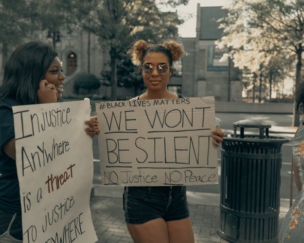 A photo of two women at a protest. One is facing the camera holding a sign that says "We WON'T be silent. No Justice No Peace" the other is facing to the side with a sign that says "injustice anywhere is a threat to justice everywhere"