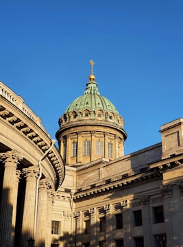 A classical-style building with ornate columns and a large dome topped with a cross. The structure is bathed in golden sunlight against a clear blue sky.