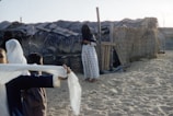 A woman stands in front of a traditional thatched hut made of wood and palm leaves, wearing a long dress with a pattern and a veil. Nearby, two children are playing on the sandy ground, one of them holding a white cloth. The setting seems to be in a rural or desert area, with clear sky visible above.
