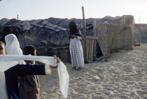 A woman stands in front of a traditional thatched hut made of wood and palm leaves, wearing a long dress with a pattern and a veil. Nearby, two children are playing on the sandy ground, one of them holding a white cloth. The setting seems to be in a rural or desert area, with clear sky visible above.