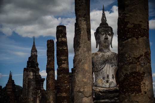 A large stone Buddha statue is surrounded by ancient stone pillars, with a backdrop of cloudy blue skies. The scene conveys a sense of historical and cultural significance.