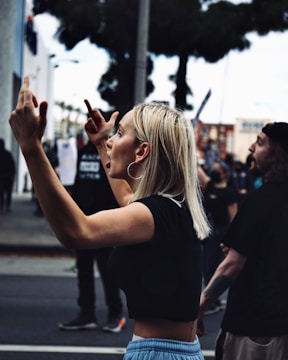 A woman with blonde hair is energetically participating in a street protest. She wears a black crop top and blue pants, raising her hands with intensity. People around her are engaged in the demonstration, with some holding signs in the background. Trees and urban buildings are visible in the surroundings.