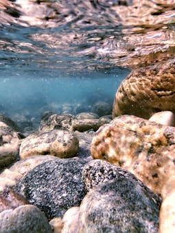 The photograph captures an underwater scene with variously sized, smooth rocks on the riverbed. The water above has gentle ripples with a light diffusing effect, creating an ethereal atmosphere. Light filters through the water, casting subtle shadows and reflections on the stones.