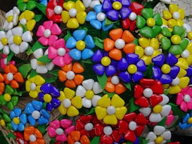 A colorful assortment of artificial flowers made of vibrant materials, featuring petals in a variety of bright colors including red, blue, yellow, orange, and pink. The flowers have noticeable white centers, and green leaves are interspersed between them. The composition creates a cheerful and lively appearance.