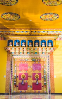 A vibrant and intricately decorated doorway, featuring rich colors and detailed patterns. The surrounding walls and ceiling are adorned with colorful murals and motifs, including circular designs that add an ornate touch. The door itself is predominantly red with golden trim and features large, ornate handles with tassels.