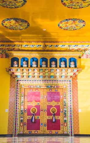 A vibrant and intricately decorated doorway, featuring rich colors and detailed patterns. The surrounding walls and ceiling are adorned with colorful murals and motifs, including circular designs that add an ornate touch. The door itself is predominantly red with golden trim and features large, ornate handles with tassels.