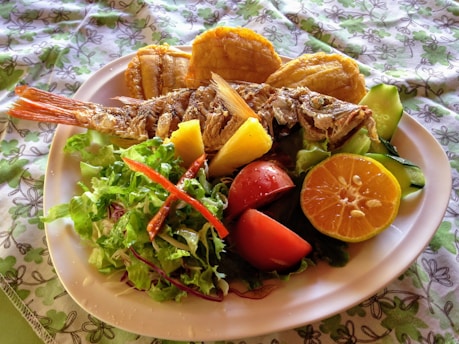 A plate filled with a variety of fresh foods, including a whole grilled fish, colorful sliced vegetables like tomatoes and cucumber, a halved orange, and a serving of fresh greens. There are also pieces of fried plantain and pineapple. The dish is presented on a white plate with a floral patterned tablecloth beneath.