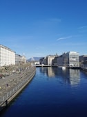 A clear waterway flows through a cityscape, flanked by modern buildings on both sides. The sky is bright blue with minimal clouds, and people are walking and sitting near the water's edge, enjoying the serene environment. In the distance, a large fountain sprays a plume of water high into the air.