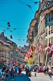 A vibrant street scene features historic buildings with intricate architecture. Numerous flags, including Swiss ones, are displayed along the street. People are walking or biking on the cobblestone path, and several parked cars are visible. The sky is clear and blue, enhancing the lively atmosphere.