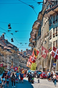 A vibrant street scene features historic buildings with intricate architecture. Numerous flags, including Swiss ones, are displayed along the street. People are walking or biking on the cobblestone path, and several parked cars are visible. The sky is clear and blue, enhancing the lively atmosphere.
